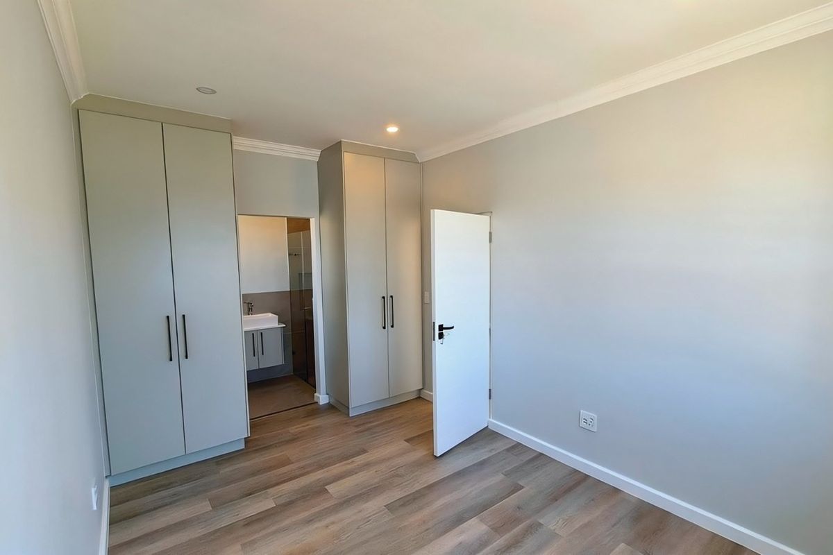 A bright, modern bedroom featuring neutral walls, laminate flooring, built-in cupboards, and an adjoining en-suite bathroom.