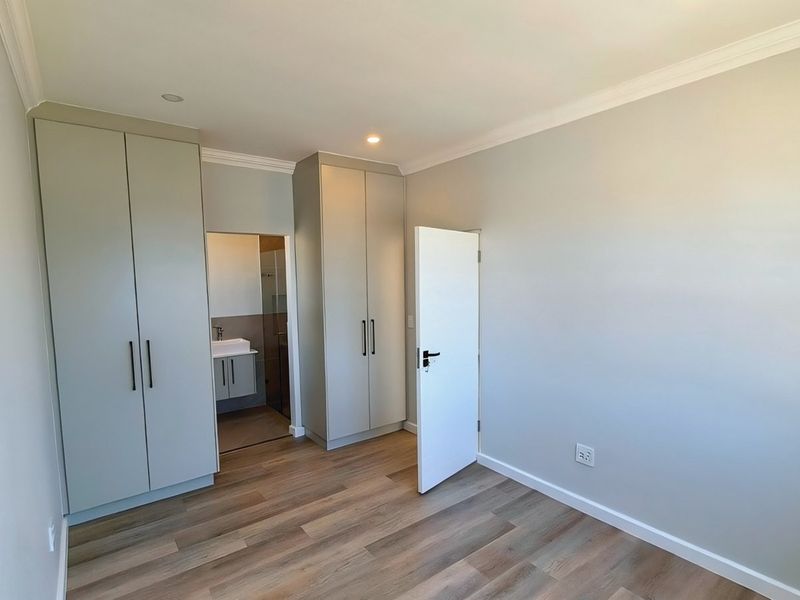 A bright, modern bedroom featuring neutral walls, laminate flooring, built-in cupboards, and an adjoining en-suite bathroom.