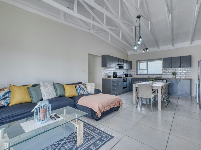 Open-plan living and kitchen area featuring high vaulted ceilings with exposed trusses, modern dark cabinetry, subway tile backsplash, and neutral floor tiling.