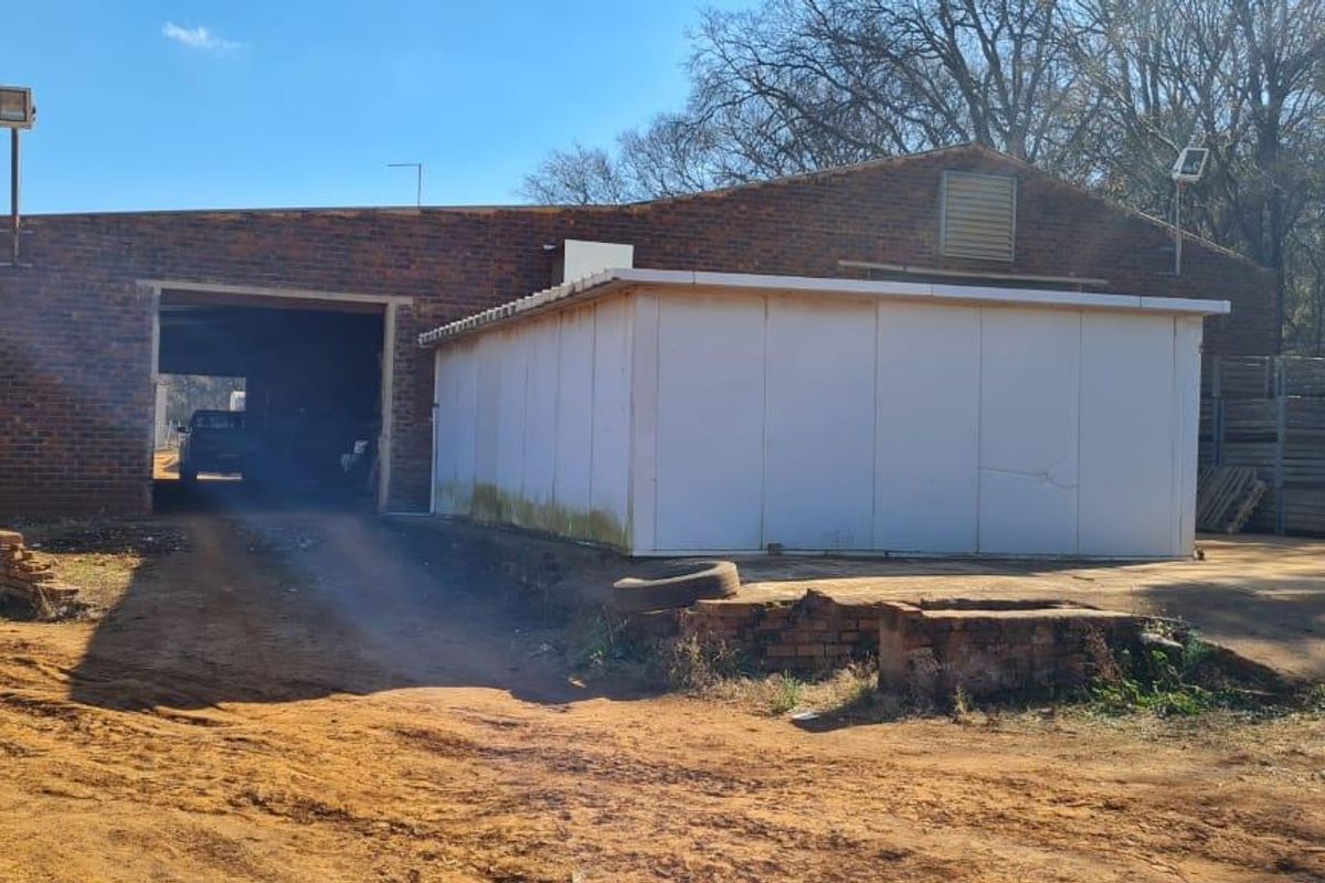 Rear view of a farm building featuring a large industrial opening, brick construction, and an attached prefabricated storage unit on a dirt yard.