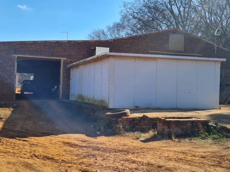 Rear view of a farm building featuring a large industrial opening, brick construction, and an attached prefabricated storage unit on a dirt yard.