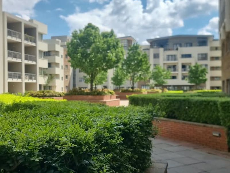 A well-maintained communal garden area featuring paved walkways, manicured hedges, and brick retaining walls between apartment blocks.