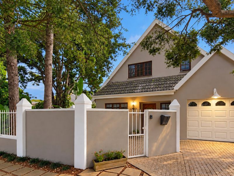 Front elevation of a well-maintained house featuring a paved driveway, garage, secure boundary wall with pedestrian gate, and mature trees.