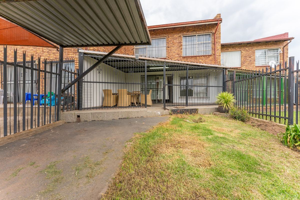 Rear view of the townhouse featuring a covered patio, brick facade, security fencing, and a paved driveway area.