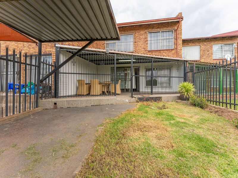 Rear view of the townhouse featuring a covered patio, brick facade, security fencing, and a paved driveway area.
