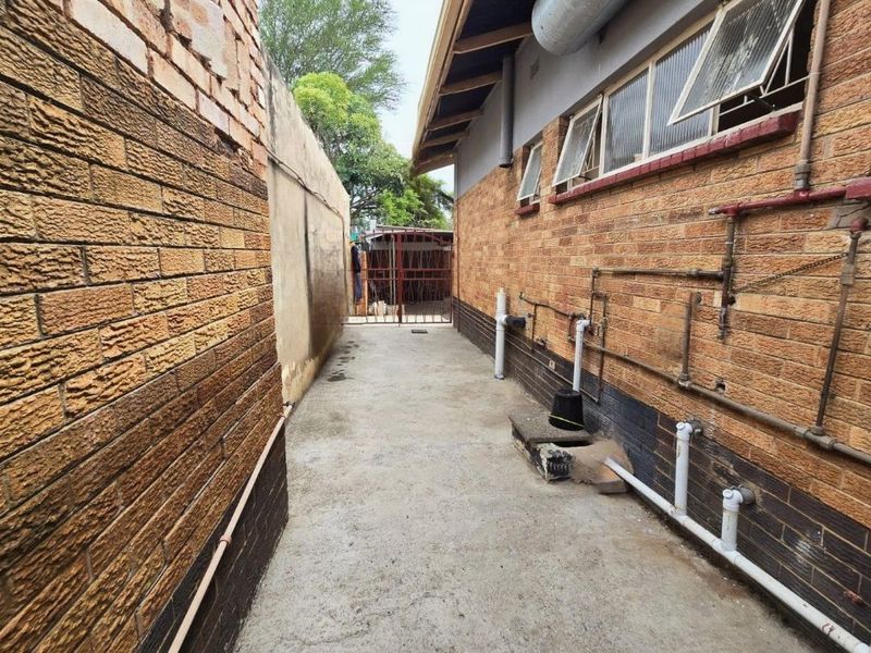A narrow side passage featuring exposed brick walls, concrete flooring, and external plumbing infrastructure leading to a rear metal security gate.