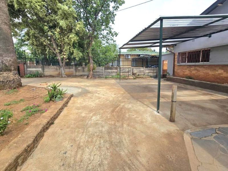 Rear exterior area featuring a covered carport, concrete driveway, and secure boundary walling with established trees.
