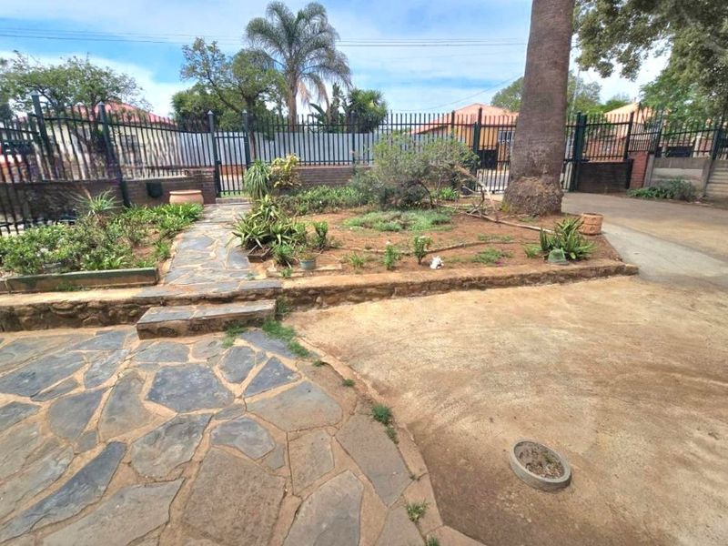 Front exterior area featuring a stone-paved walkway, driveway, metal perimeter fencing, and established garden beds with a large tree.