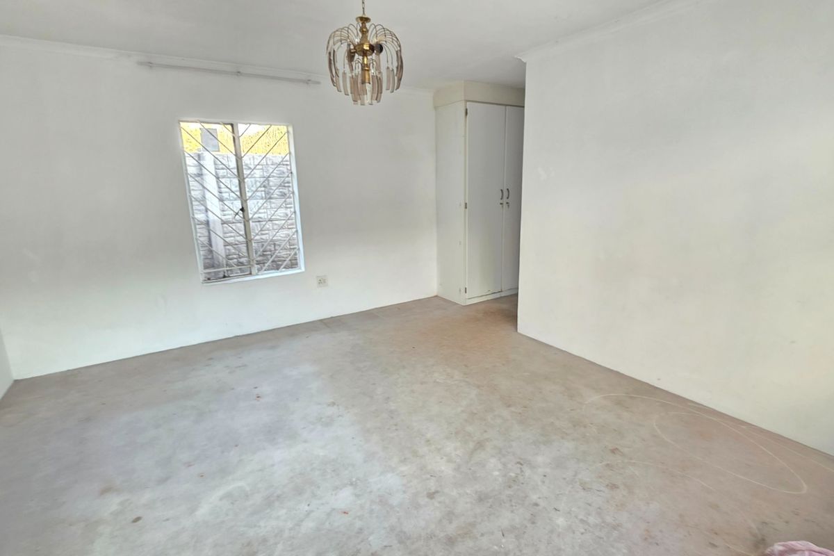 An empty bedroom featuring a built-in cupboard, security bars on the window, and a central light fixture, awaiting new flooring and finishing touches.