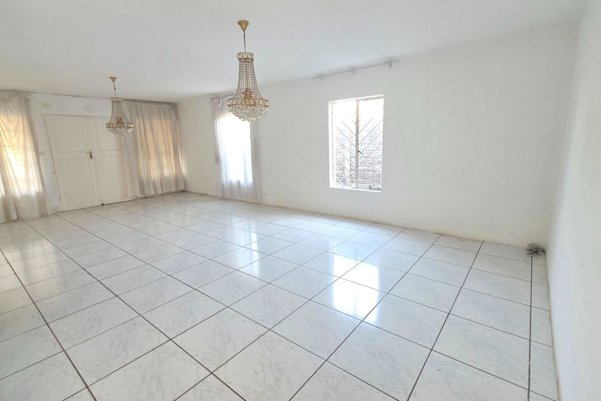 A spacious, empty living room featuring white ceramic floor tiles, crystal chandeliers, and natural light through windows with security bars.