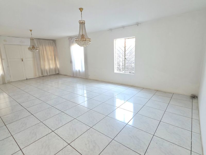A spacious, empty living room featuring white ceramic floor tiles, crystal chandeliers, and natural light through windows with security bars.