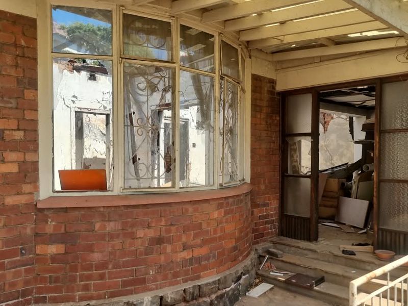 A dilapidated covered patio area featuring a brick facade, bay window with security bars, and visible structural damage to the interior and exterior finishes.