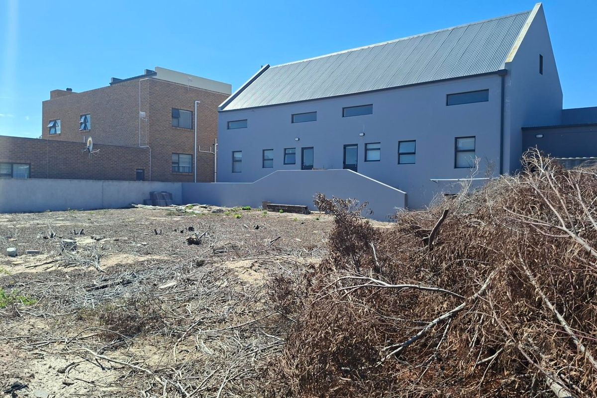 A view of a vacant residential plot featuring a boundary wall and a view of the adjacent modern grey building with a metal roof.
