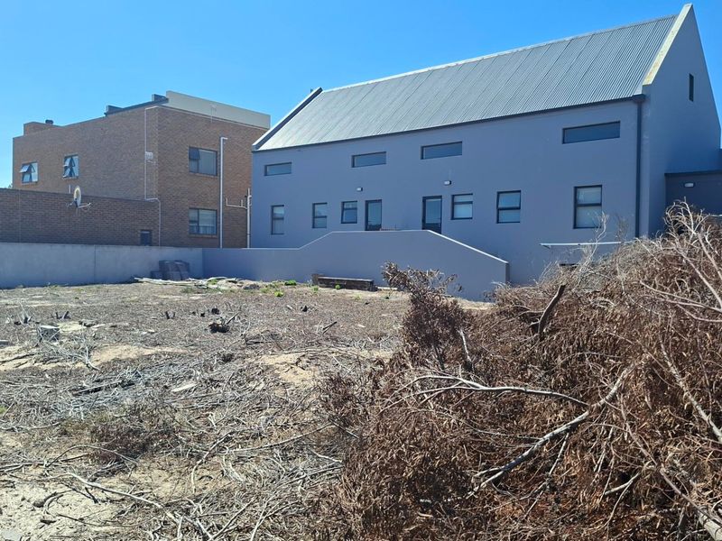 A view of a vacant residential plot featuring a boundary wall and a view of the adjacent modern grey building with a metal roof.
