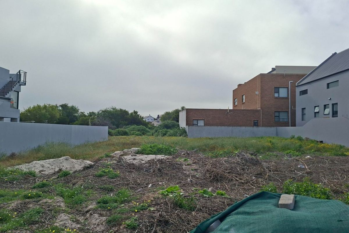 A vacant residential plot situated between existing residential properties, featuring boundary walls and natural ground cover.