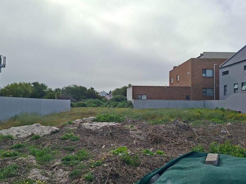 A vacant residential plot situated between existing residential properties, featuring boundary walls and natural ground cover.
