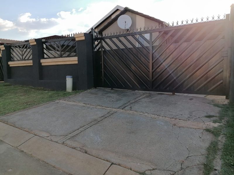 Front view of the property featuring a secure metal driveway gate, perimeter wall, and a concrete driveway with visible surface cracking.