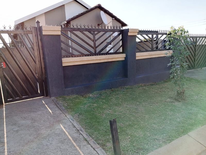 Front view of the property featuring a secure perimeter wall with metal fencing, a matching pedestrian gate, and a well-maintained front lawn.