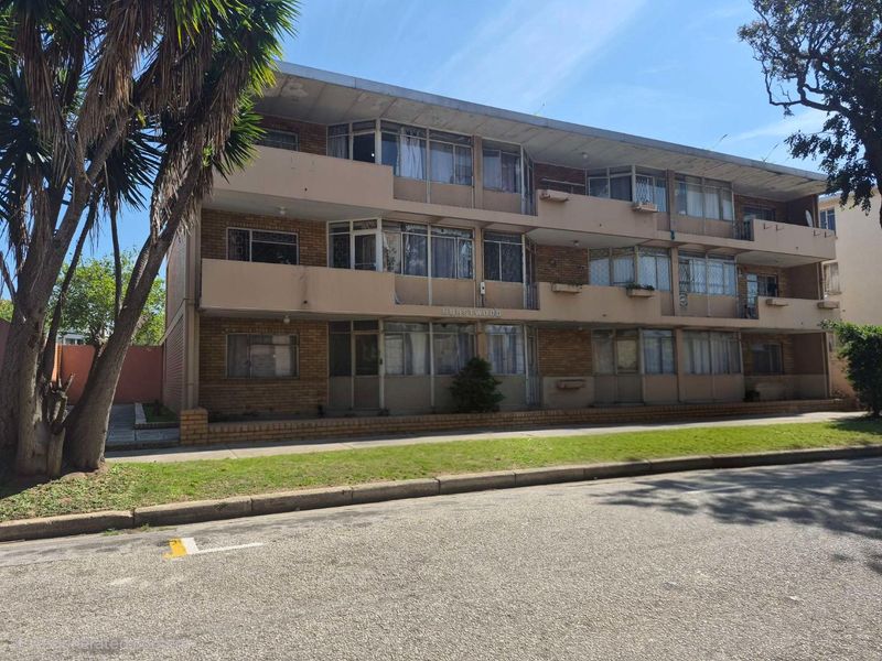 A three-story apartment building facade featuring brick construction, private balconies, and a concrete roof overhang, situated along a street.