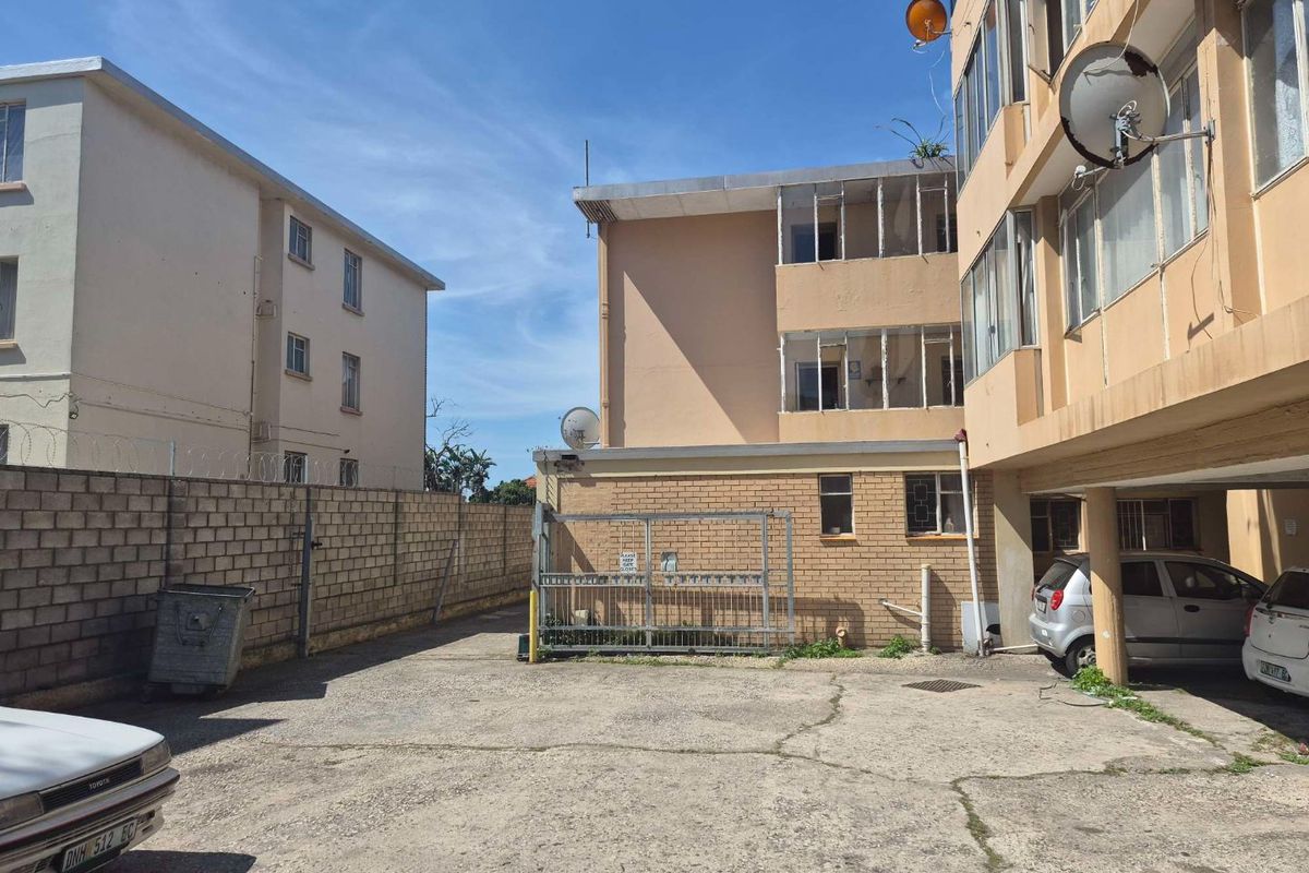 Rear exterior view of the apartment complex featuring a concrete parking area, brick boundary wall, and multi-story building facade with enclosed balconies.