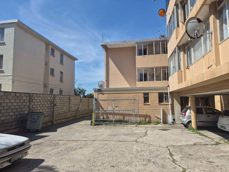 Rear exterior view of the apartment complex featuring a concrete parking area, brick boundary wall, and multi-story building facade with enclosed balconies.
