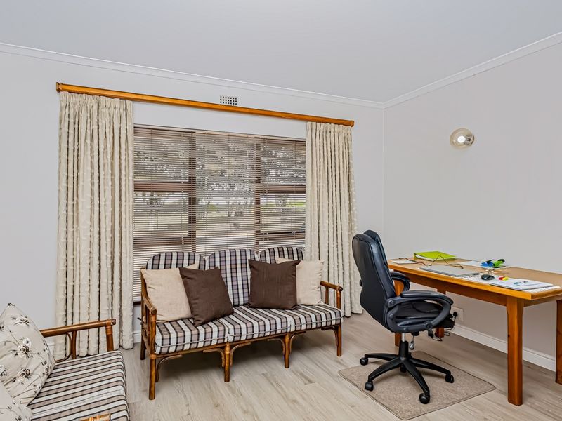 A functional home office space featuring light-toned laminate flooring, neutral walls, and large windows fitted with venetian blinds and curtains.