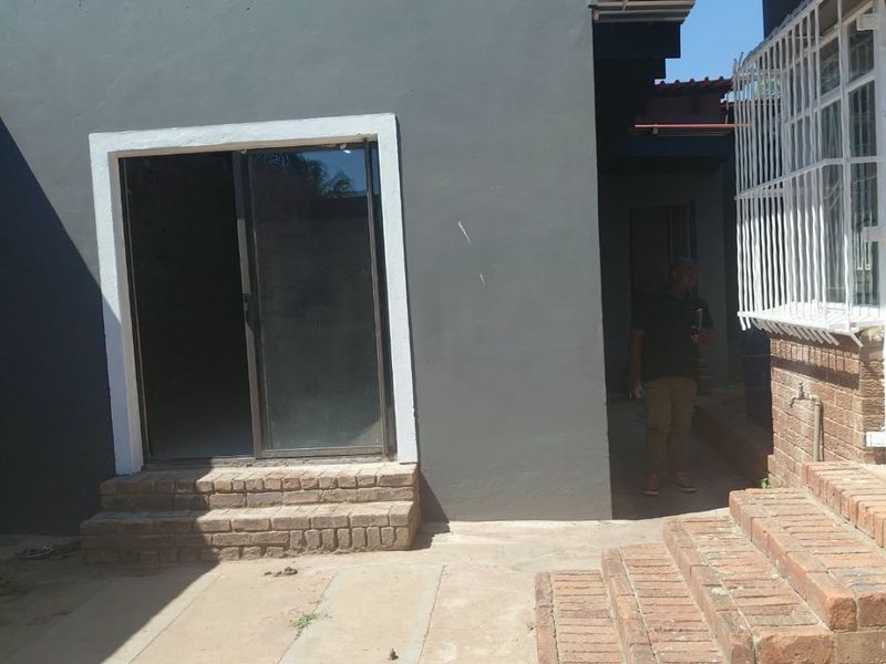 Rear exterior view showing a grey painted wall, sliding glass door access, brick steps, and concrete paving with security bars on the adjacent window.