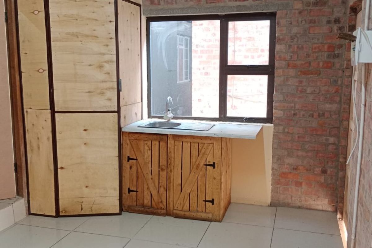 A compact kitchen area featuring exposed brick walls, ceramic tiled flooring, and basic wooden cabinetry with a single basin sink.