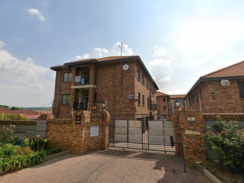 Exterior view of a multi-story face brick apartment complex featuring secure gated access, paved driveway, and perimeter electric fencing.