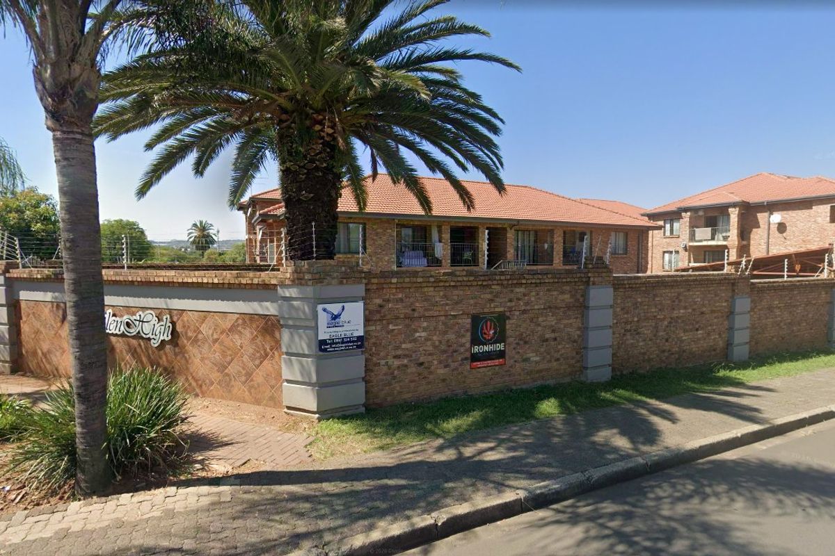 Street view of the apartment complex entrance featuring a brick perimeter wall, electric fencing, and mature palm tree landscaping.