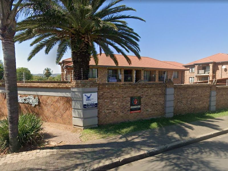 Street view of the apartment complex entrance featuring a brick perimeter wall, electric fencing, and mature palm tree landscaping.