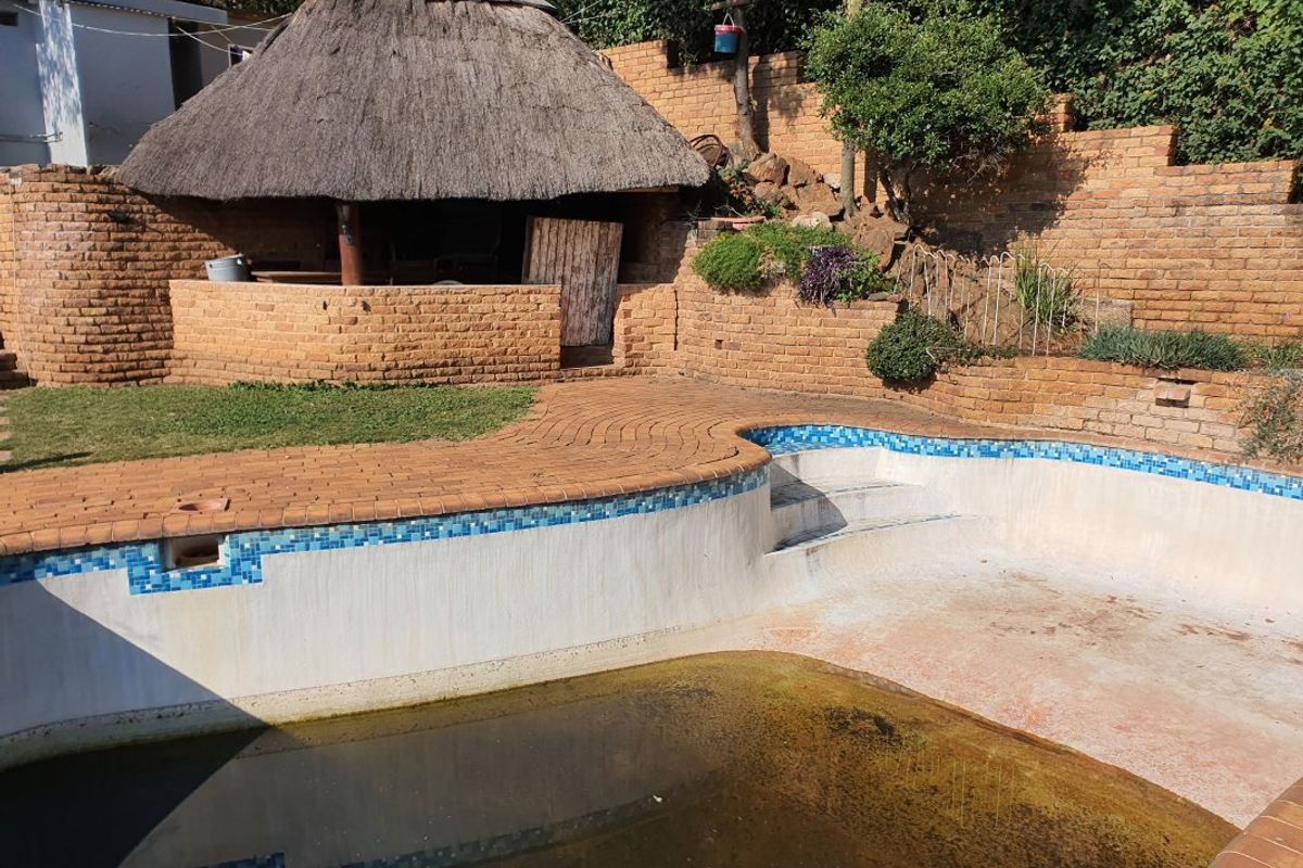 Outdoor pool area featuring a thatched lapa, brick retaining walls, and paved surround, currently requiring significant maintenance and pool restoration.