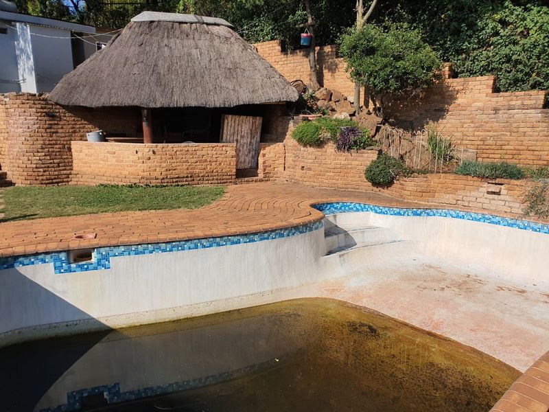 Outdoor pool area featuring a thatched lapa, brick retaining walls, and paved surround, currently requiring significant maintenance and pool restoration.