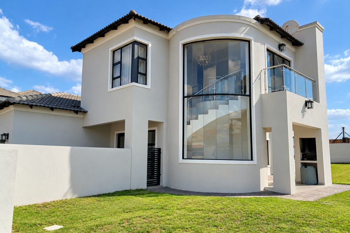 Modern double-story home exterior featuring a striking curved glass facade, balcony with glass balustrade, and a covered patio area with built-in braai.