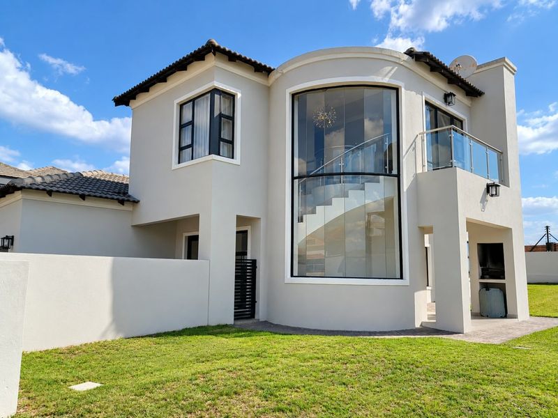 Modern double-story home exterior featuring a striking curved glass facade, balcony with glass balustrade, and a covered patio area with built-in braai.