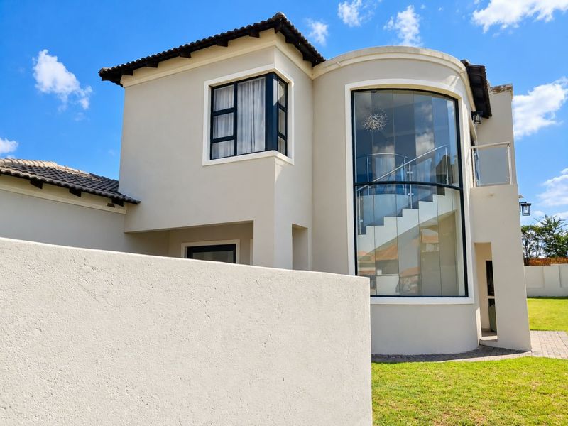 A modern double-story home exterior featuring a striking curved glass facade, a prominent staircase, and a well-maintained garden with a boundary wall.