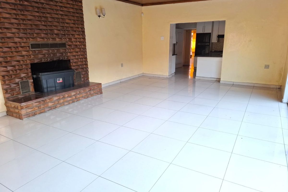 Spacious living area featuring a prominent brick fireplace, tiled flooring, and a timber ceiling, with an open-plan flow into the adjacent kitchen area.