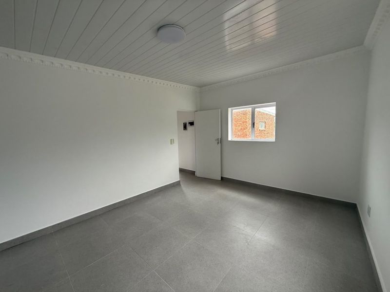 A clean, empty bedroom featuring neutral grey floor tiles, a white tongue and groove ceiling with decorative cornicing, and a single window.
