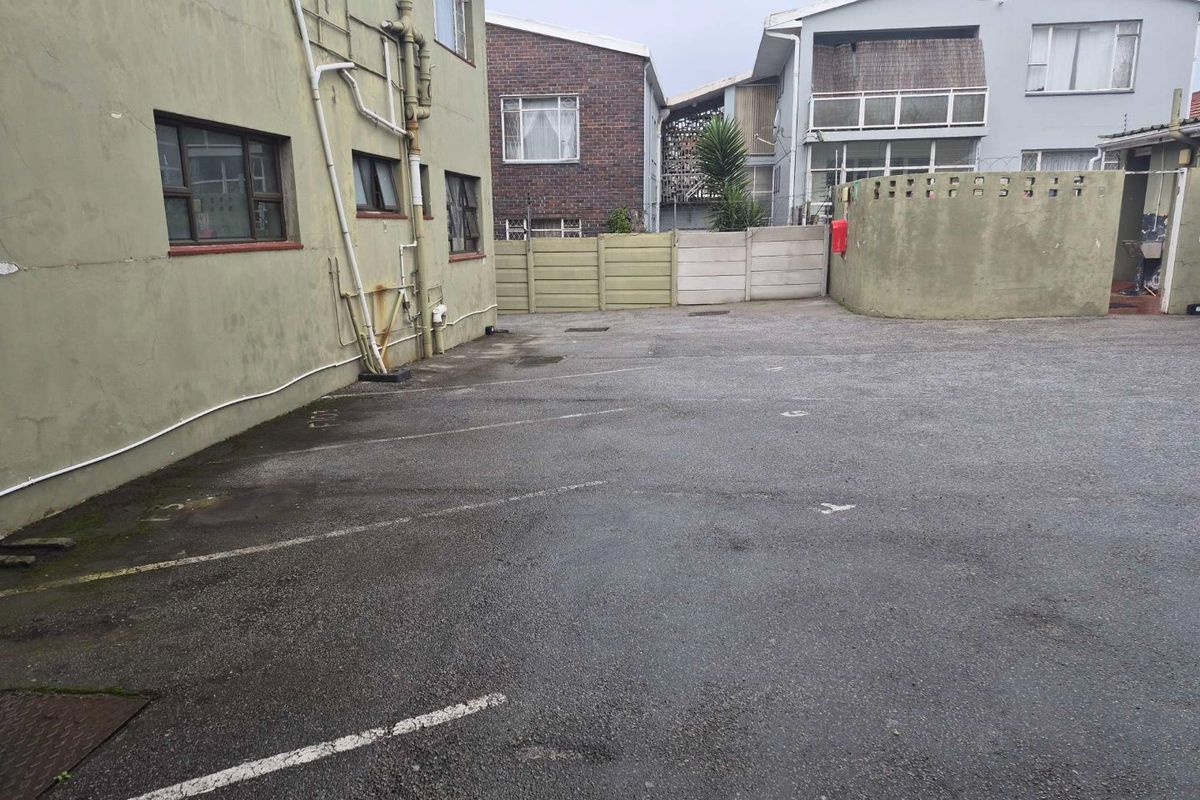Rear exterior view of the apartment building showing an asphalt parking area, boundary walls, and exposed utility piping.