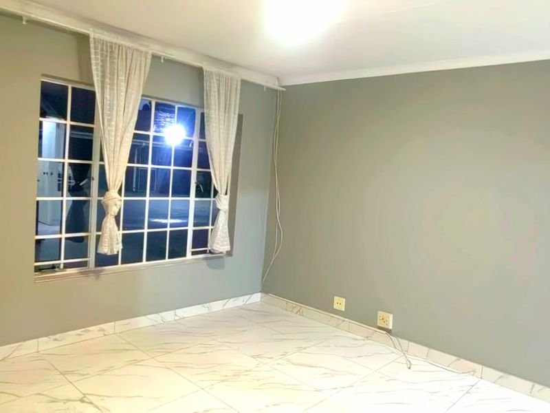 A bright, empty bedroom featuring modern marble-look tiled flooring, neutral grey walls, and a large grid-pattern window allowing for natural light.