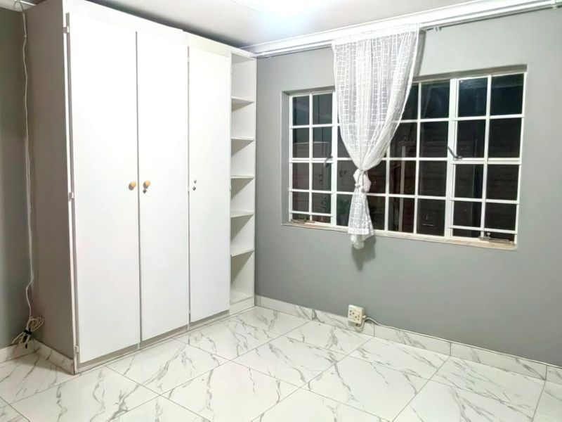 A clean, empty bedroom featuring modern marble-look floor tiles, a built-in wardrobe, and a window with security bars.