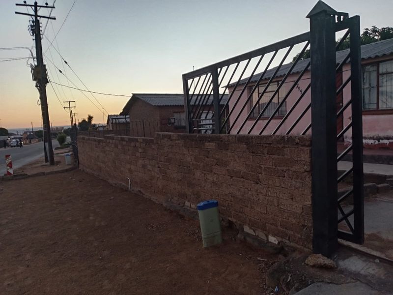 Front view of the property featuring a brick boundary wall and metal security gate, with visible exterior wall finishes and security bars on windows.