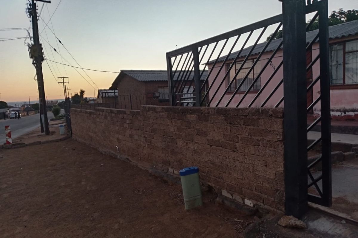 Front view of the property featuring a brick boundary wall and metal security gate, with visible exterior wall finishes and security bars on windows.