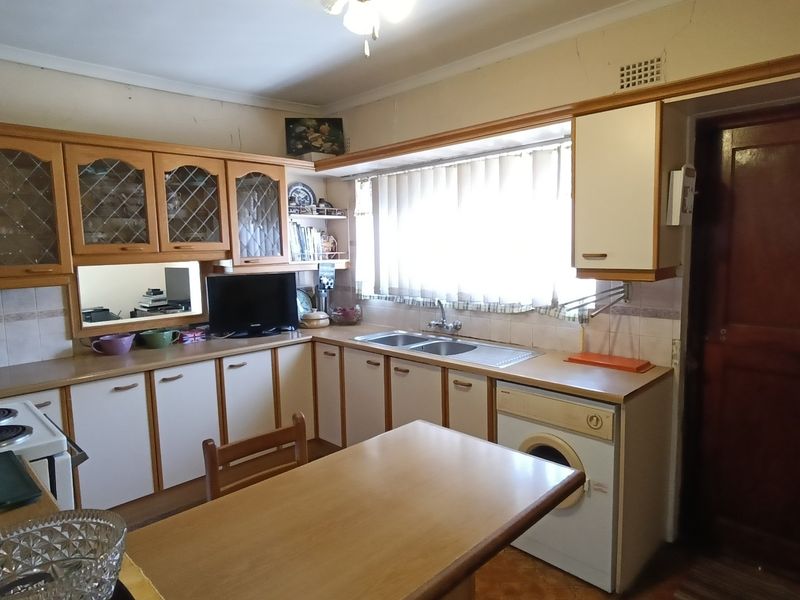 A dated kitchen featuring laminate cabinetry, a double bowl sink, tiled backsplash, and integrated laundry facilities, requiring cosmetic updates to modernize the space.