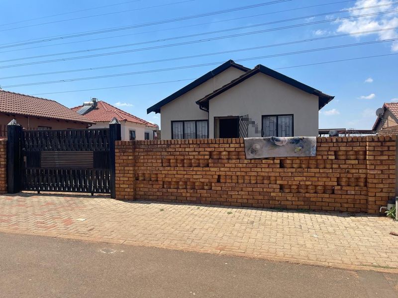 Front view of a single-story house featuring a brick boundary wall, metal driveway gate, and paved entrance area.