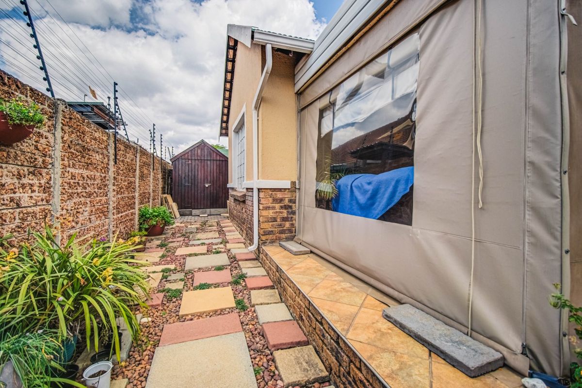 Rear exterior view featuring a paved walkway, brick boundary wall with electric fencing, and a covered patio area with canvas enclosures.