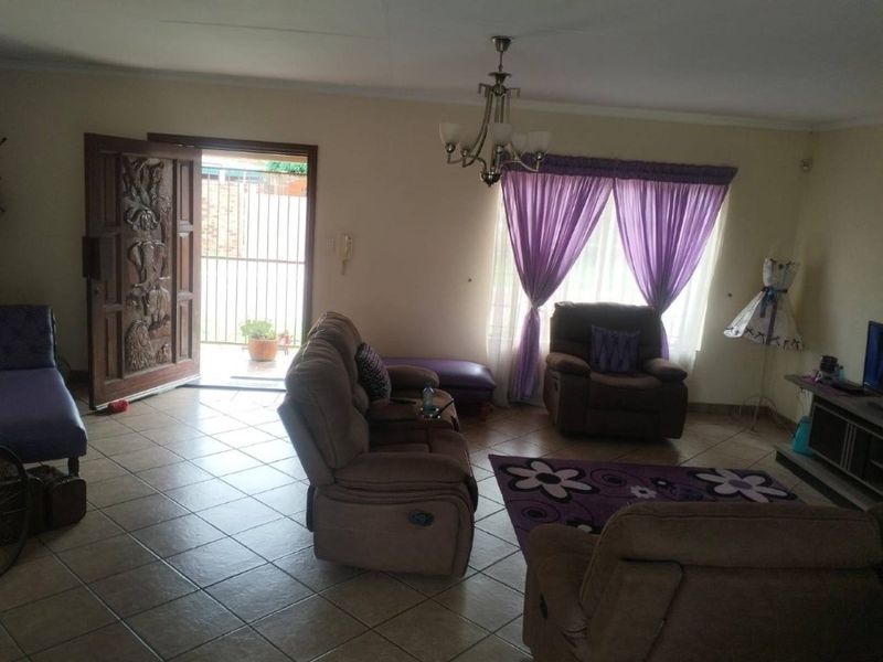 A spacious living room featuring tiled flooring, an ornate wooden entrance door, and a central chandelier, providing a functional and welcoming living space.
