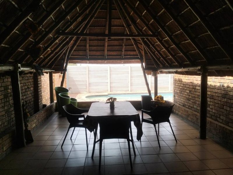 A covered outdoor patio area featuring a traditional thatched roof, exposed brick walls, and tiled flooring, overlooking a private swimming pool.