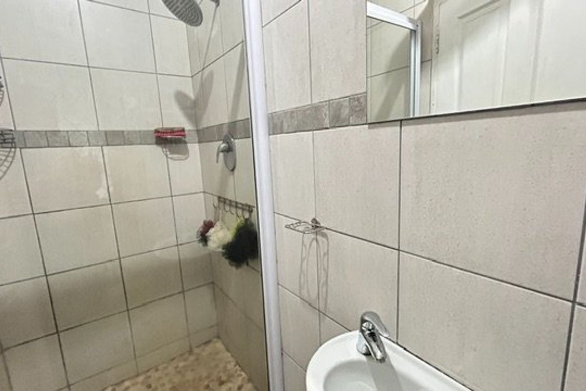 A functional bathroom featuring a tiled shower enclosure, pedestal basin, and wall-mounted mirror, presenting a standard layout with dated finishes.