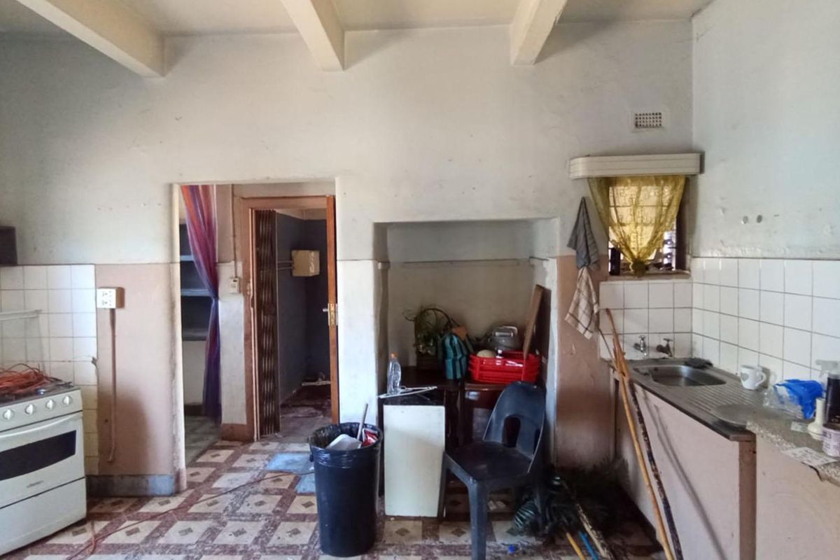 A dated kitchen featuring a freestanding stove, stainless steel sink, tiled splashbacks, and exposed ceiling beams, requiring significant renovation and cosmetic updates.